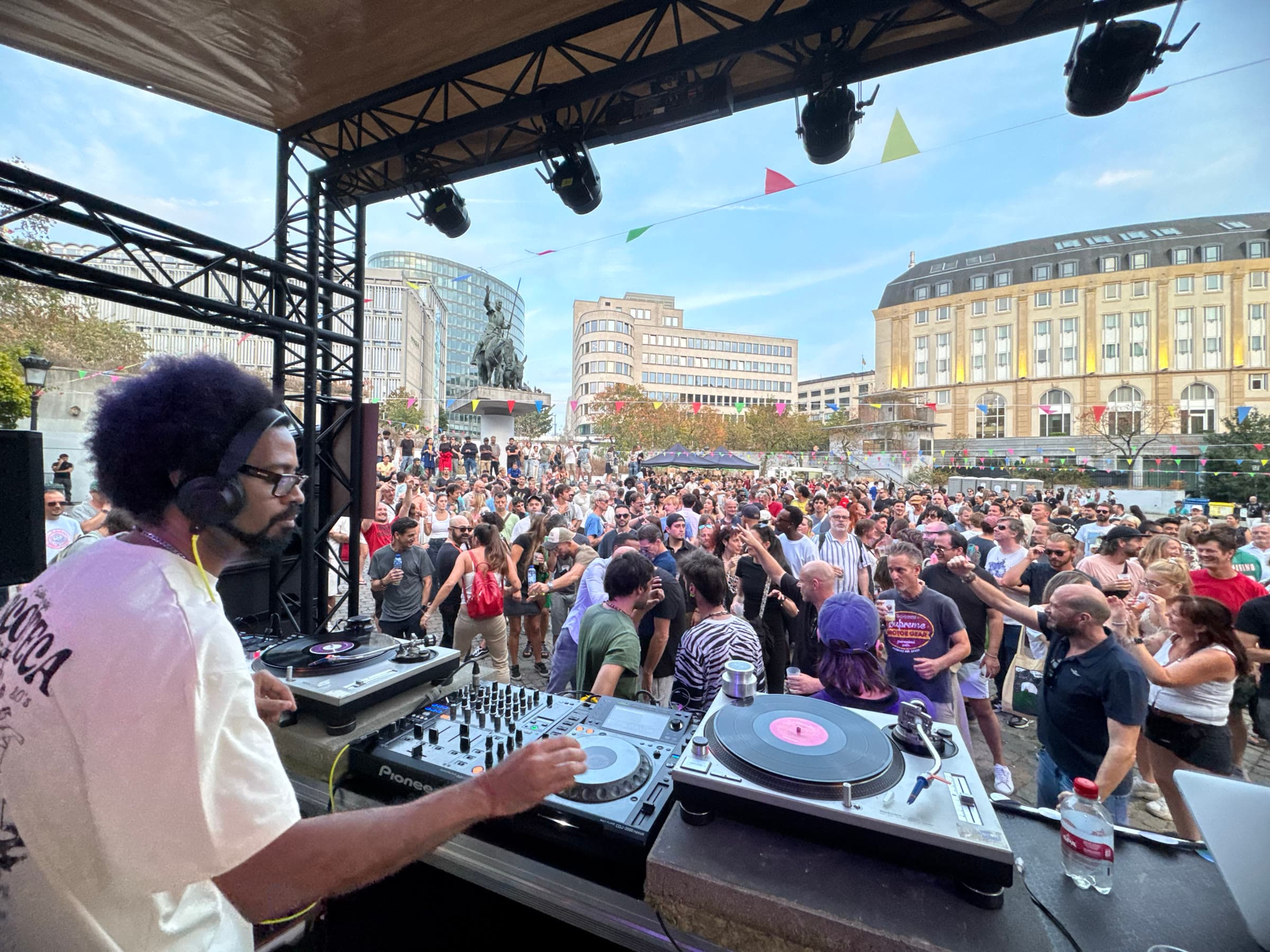 A DJ at the decks on Place d'Espagne, facing a packed square in late afternoon sun.