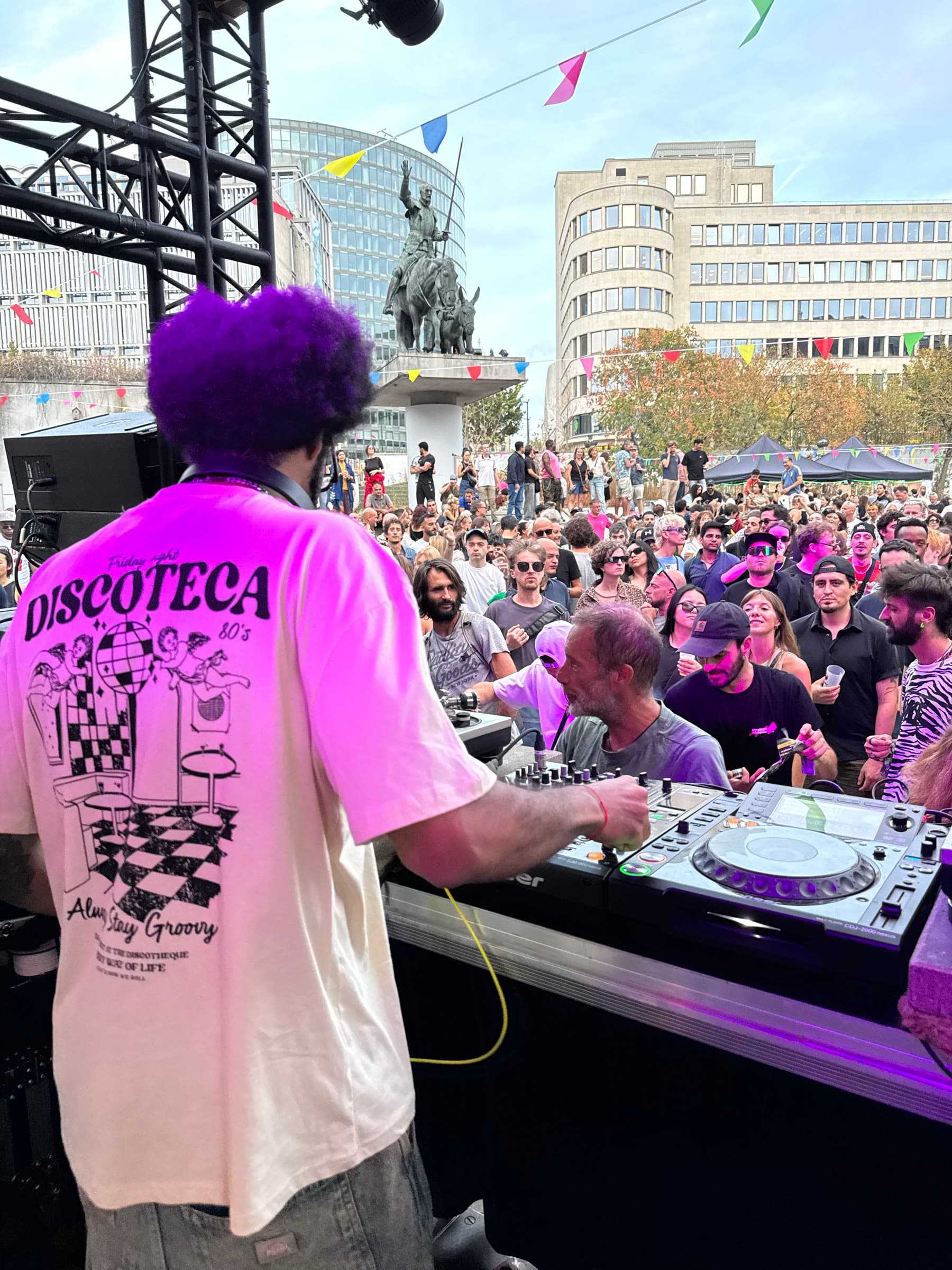 Seen from behind the decks: a DJ in a printed shirt above a crowd filling the square.
